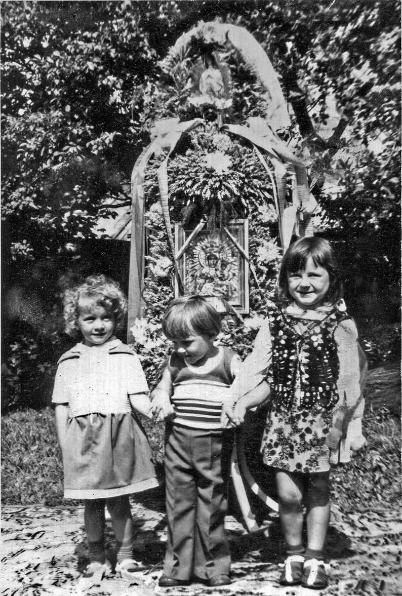 trójka małych dzieci pozuje do zdjęcia trzymając się za ręce. Za nimi wieniec dożynkowy. Zdjęcie czarnobiałe